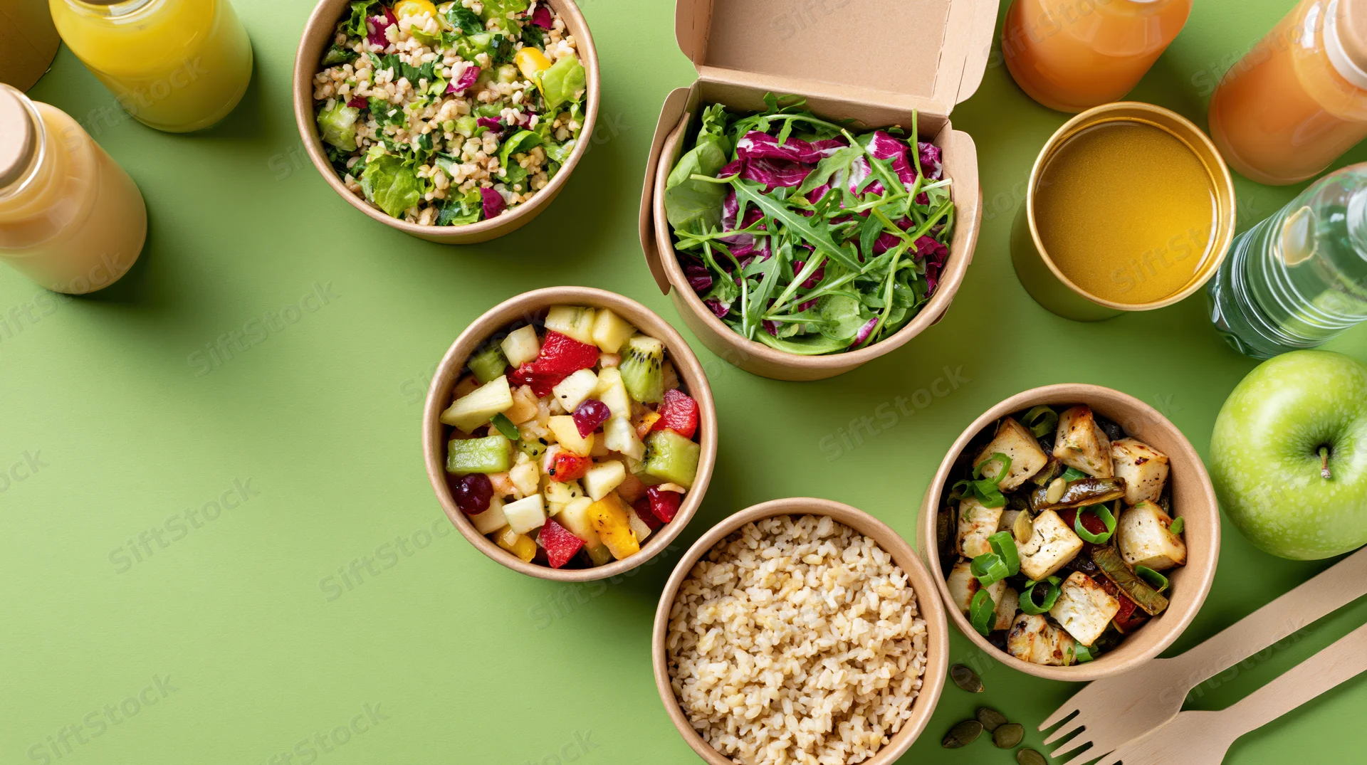 Overhead flat lay of healthy meal prep containers with salads, fruit, grains, and drinks on green surface