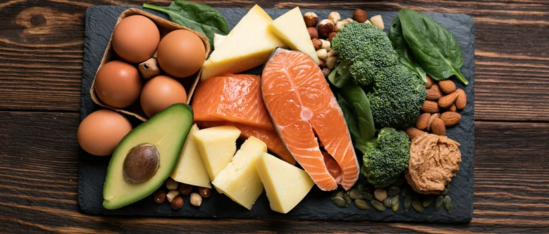 Overhead flat lay of keto diet foods arranged on black slate board with salmon, eggs, avocado, cheese, broccoli, nuts and spinach on wooden table