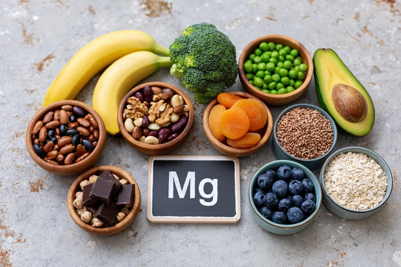 Overhead flat lay of magnesium-rich foods arranged on a gray surface with bowls of fruits, vegetables, nuts, seeds, and grains surrounding a chalkboard sign labeled Mg