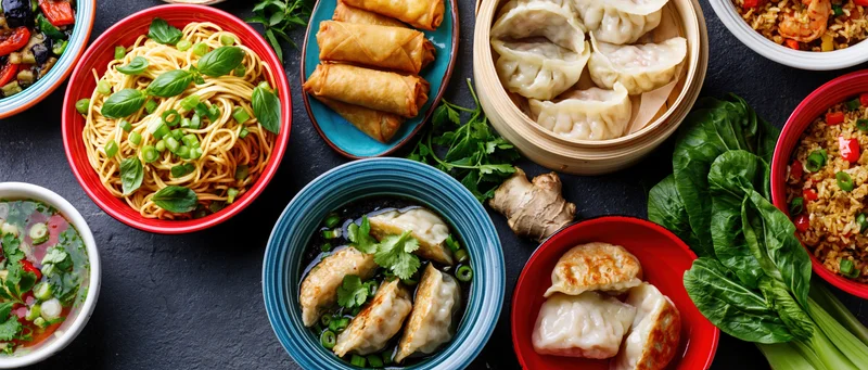 Overhead flat lay of various asian dishes including dumplings, noodles, spring rolls, and fried rice arranged on a dark surface with fresh herbs and ginger