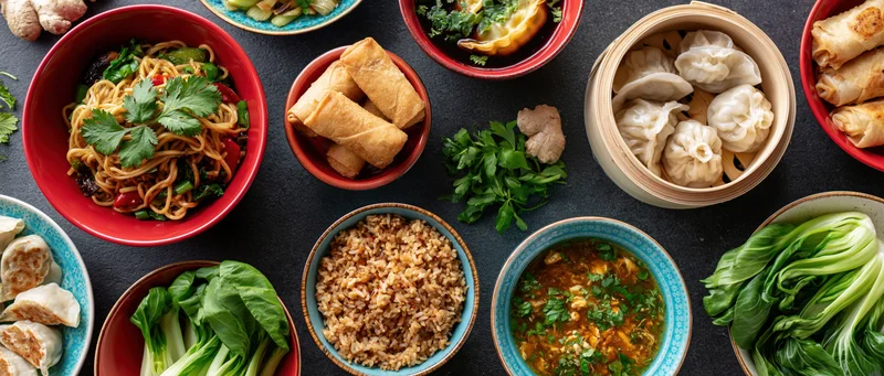 Overhead flat lay of multiple asian dishes arranged on dark surface with bowls of noodles, rice, dumplings, spring rolls, bok choy, and garnishes