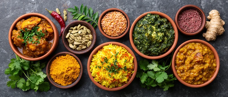 Overhead flat lay of multiple Indian dishes arranged on a dark stone surface with bowls of curry, rice, lentils, spices, and fresh herbs
