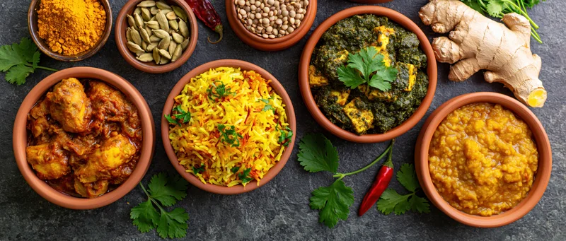 Overhead flat lay of multiple indian dishes arranged on dark stone surface with clay bowls and fresh herbs