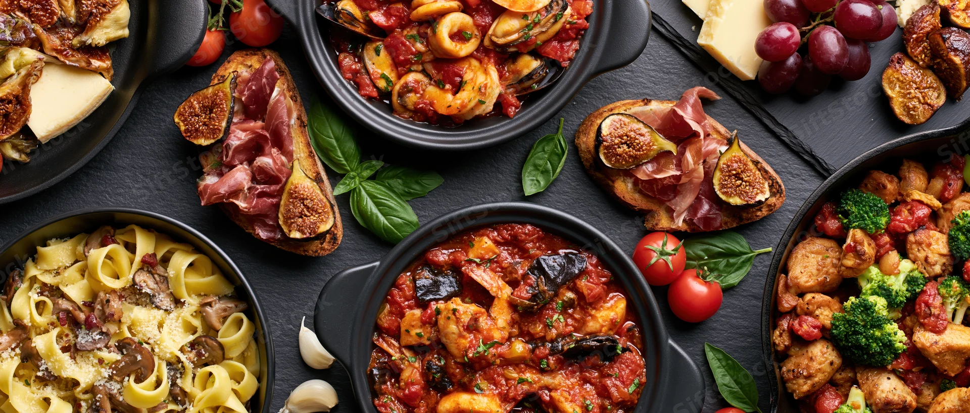 Overhead flat lay of multiple italian dishes arranged on a black slate surface with pasta, seafood, meat, vegetables, cheese, figs, prosciutto, tomatoes, basil, garlic, grapes, and bread