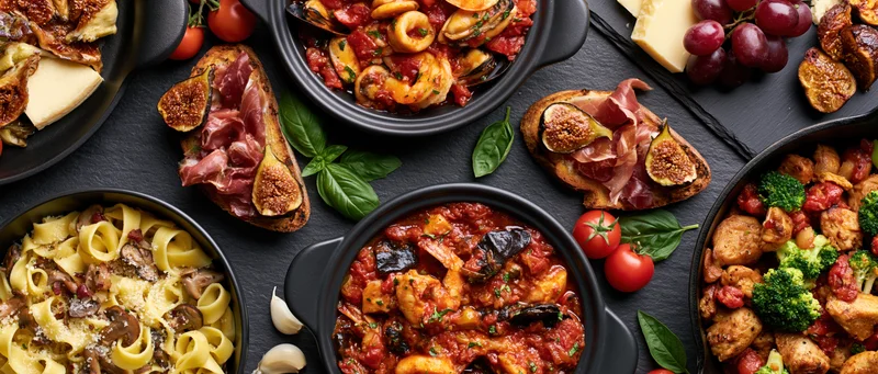 Overhead flat lay of multiple italian dishes arranged on a black slate surface with pasta, seafood, meat, vegetables, cheese, figs, prosciutto, tomatoes, basil, garlic, grapes, and bread