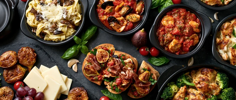 Overhead flat lay of multiple italian dishes arranged on a dark slate surface with cheese, figs, basil and tomatoes