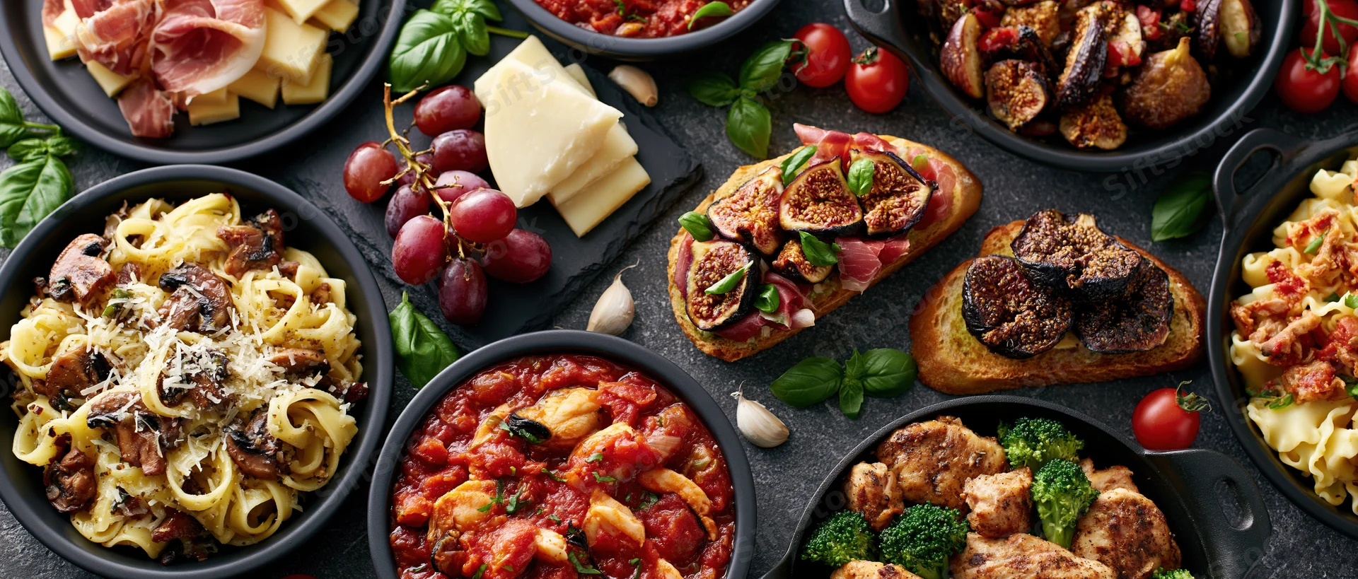 Overhead flat lay of multiple italian dishes arranged on a dark surface with pasta, cheese, figs, grapes, and vegetables