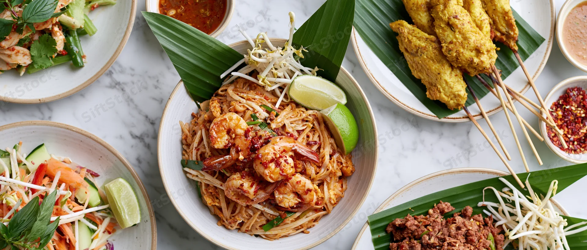 Overhead flat lay of multiple thai dishes including padthai with shrimp, satay skewers, and salads arranged on a white marble table