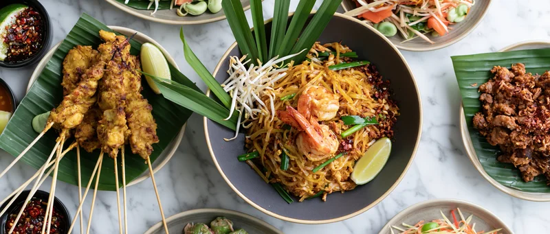 Overhead flat lay of multiple thai dishes arranged on a marble surface with satay, pad thai, and garnishes