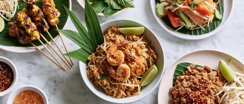 Overhead flat lay of multiple thai dishes including padthai, satay, and papaya salad arranged on a white marble table with lime wedges and herbs