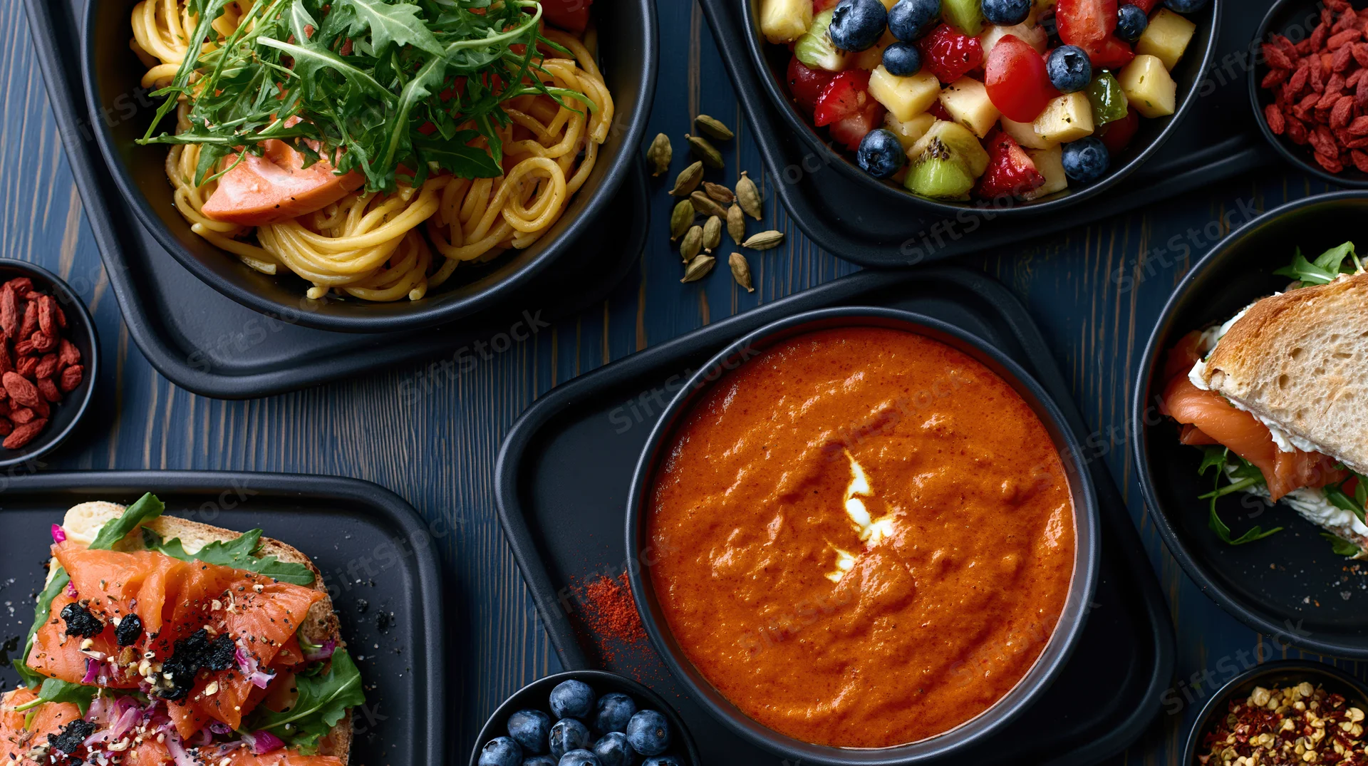 Overhead flat lay of multiple black trays and bowls with pasta, soup, sandwiches, fruit salad, and garnishes on a dark wooden table