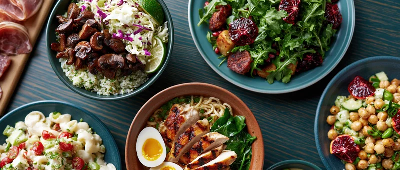 Overhead flat lay of multiple dishes including salads, pasta, rice and grilled chicken arranged on a dark wooden table