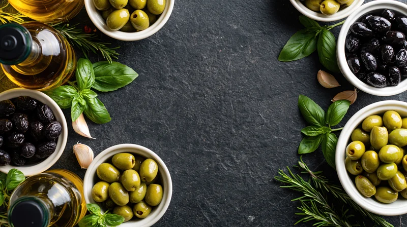 Overhead flat lay of green and black olives in white ceramic bowls with olive oil bottles, basil, garlic, and rosemary arranged on dark slate surface
