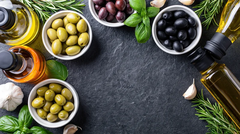 Overhead flat lay of olive oil bottles, ceramic bowls with olives, garlic, basil and rosemary arranged on dark slate surface