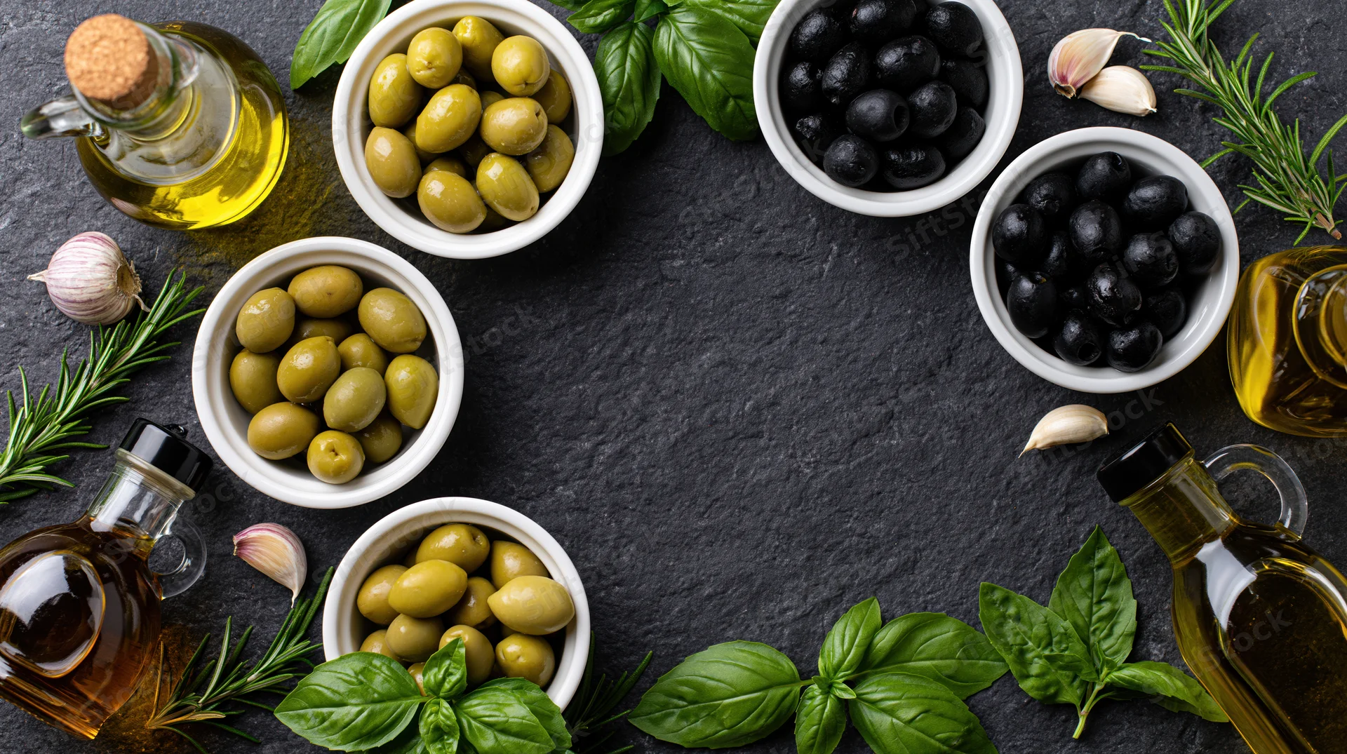 Overhead flat lay of olive oil bottles and ceramic bowls with green and black olives, garlic, rosemary, and basil arranged on dark stone surface