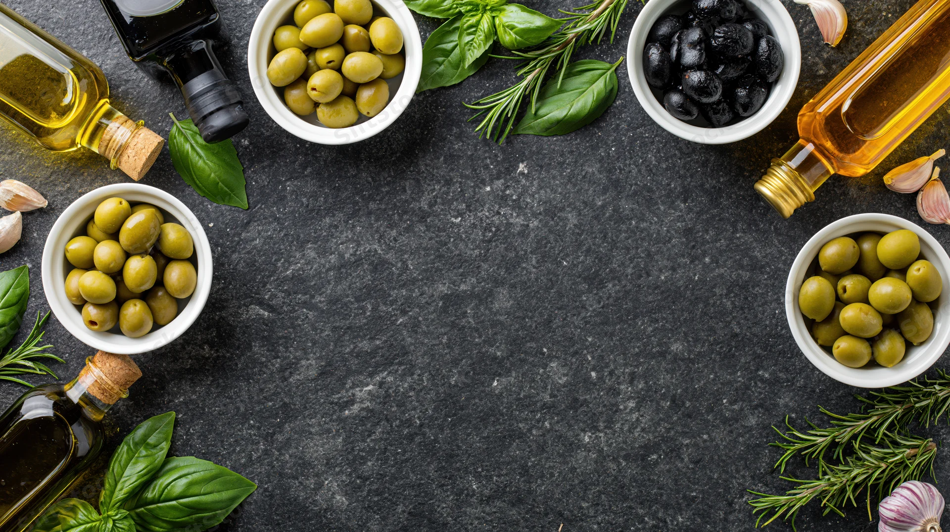 Overhead flat lay of olive oil bottles, ceramic bowls with green and black olives, garlic, basil and rosemary arranged around dark stone surface with central copyspace
