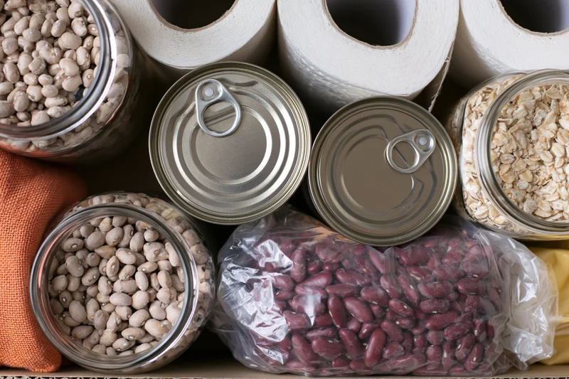 Overhead flat lay of pantry staples including canned beans, jars of oats and white beans, paper towels, and plastic bags on a cardboard surface