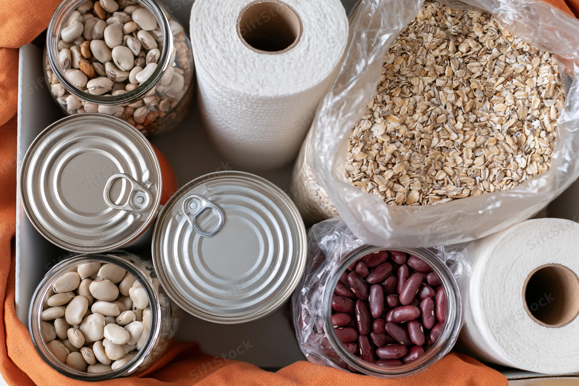 Overhead flat lay of pantry staples and household supplies arranged in a tray with jars of beans, oats in a bag, canned goods, and paper towels on an orange cloth