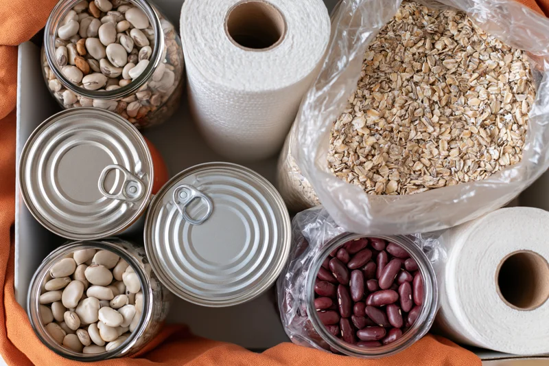 Overhead flat lay of pantry staples and household supplies arranged in a tray with jars of beans, oats in a bag, canned goods, and paper towels on an orange cloth