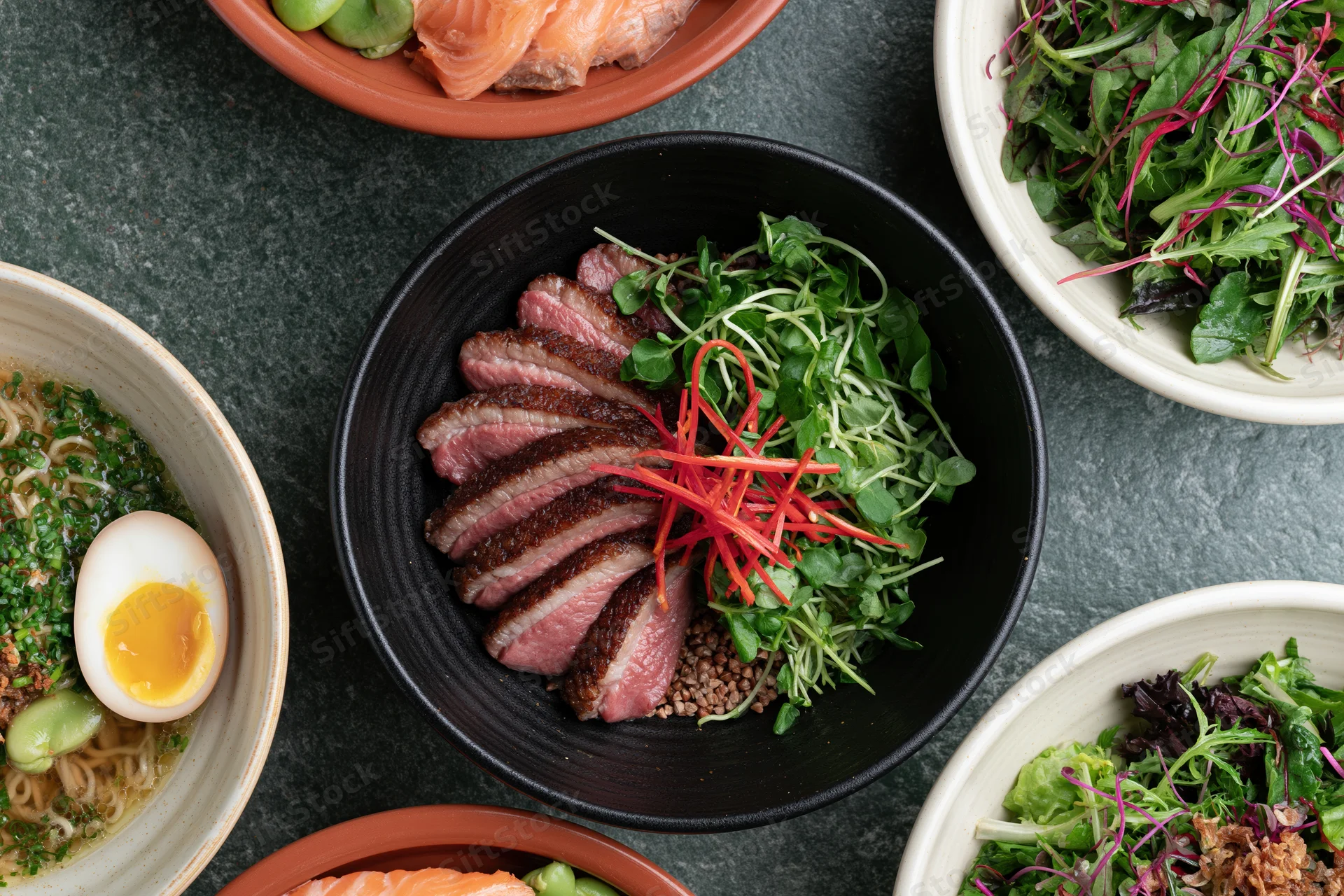 Overhead flat lay of six Asian fusion dishes arranged on dark surface with bowls of ramen, beef salad, salmon, and greens