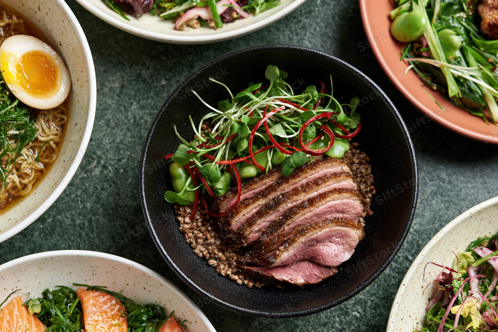 Overhead flat lay of multiple Asian fusion dishes arranged on dark surface with bowls of meat, salad, noodles and egg