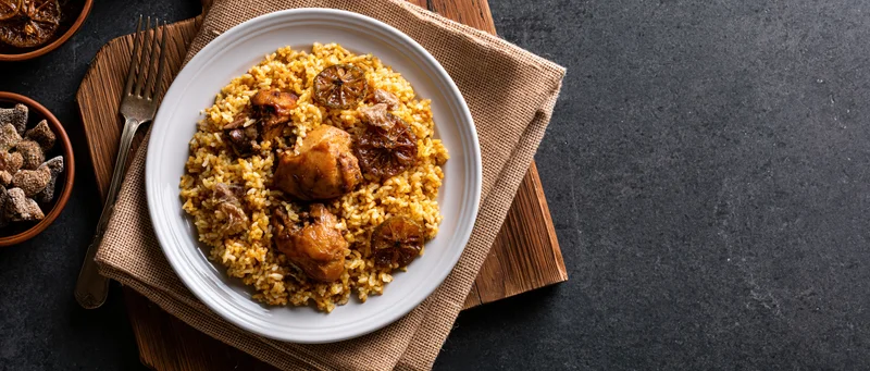 Spiced rice pilaf with braised chicken served on a white plate atop a burlap napkin and wooden board, viewed from above on a dark textured surface