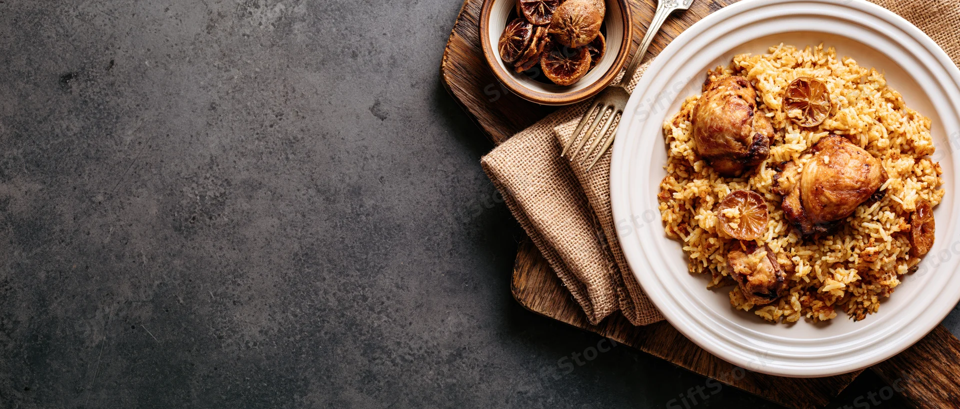 Spiced rice pilaf with braised chicken on a white plate, garnished with dried citrus slices, arranged on a wooden tray with a fork and burlap napkin on a dark surface