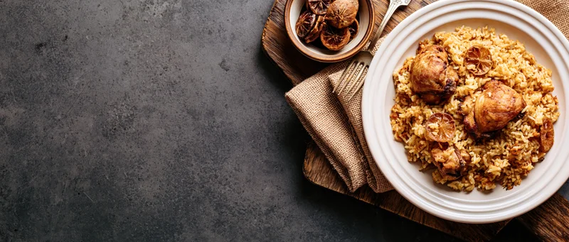 Spiced rice pilaf with braised chicken on a white plate, garnished with dried citrus slices, arranged on a wooden tray with a fork and burlap napkin on a dark surface
