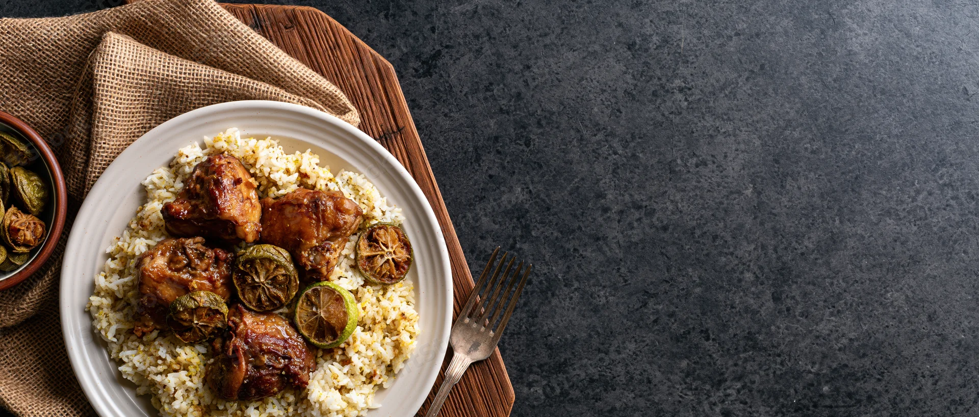 Braised chicken on spiced rice pilaf served on white plate with lime garnish on wooden tray with burlap napkin and fork on dark stone surface