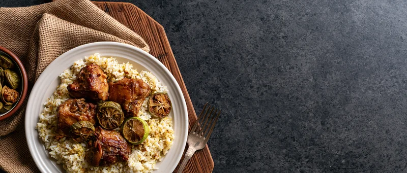Braised chicken on spiced rice pilaf served on white plate with lime garnish on wooden tray with burlap napkin and fork on dark stone surface