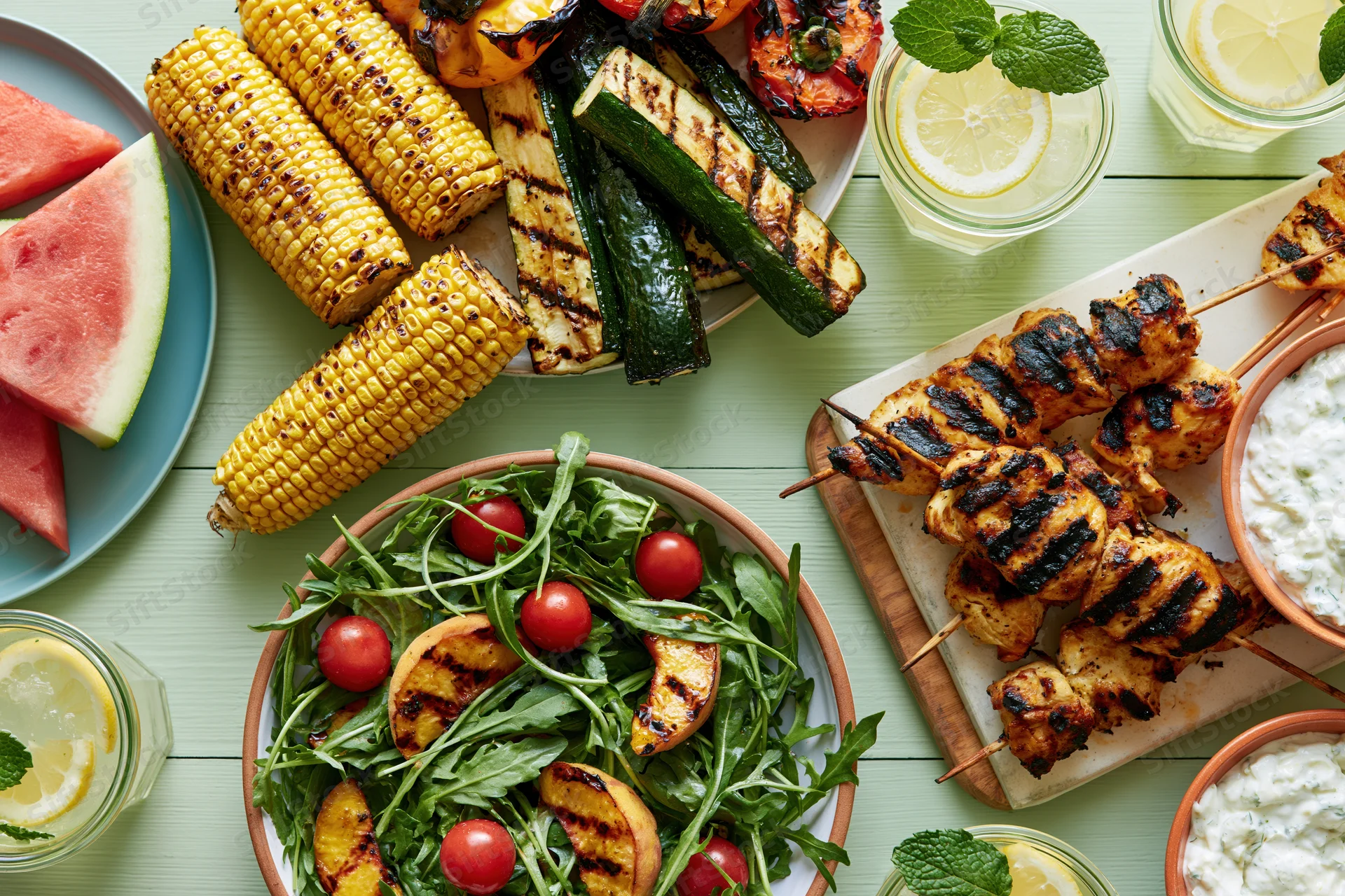 Overhead view of grilled summer barbecue spread with corn, chicken skewers, vegetables, salad, watermelon, and lemon drinks on a pale green wooden table