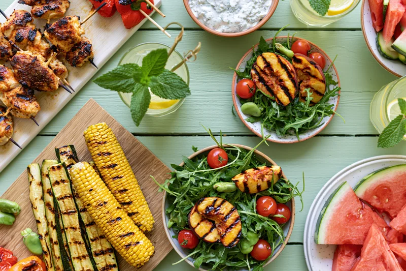 Grilled chicken and vegetables with watermelon and salads arranged on a pale green wooden table for a summer barbecue meal