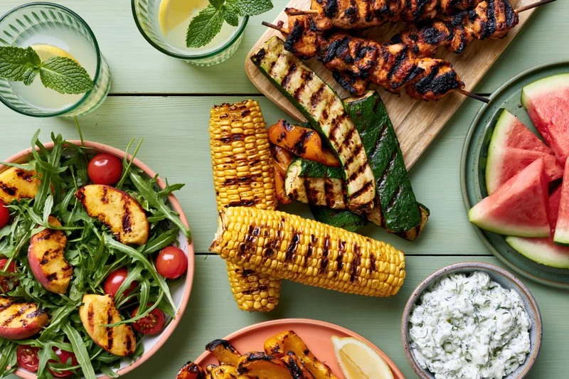 Grilled vegetables and corn with watermelon and salad arranged on a pale green wooden table for a summer barbecue