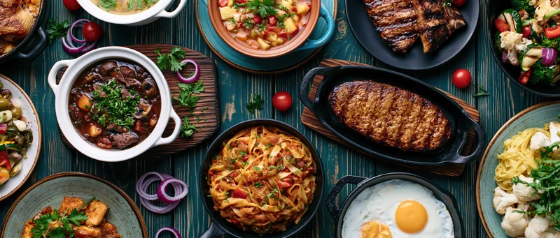 Overhead flat lay of various restaurant dishes arranged on a wooden table with garnishes and servingware