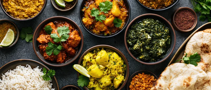 Overhead view of indian dishes arranged on dark concrete surface with bowls of curry, rice, naan, and garnishes