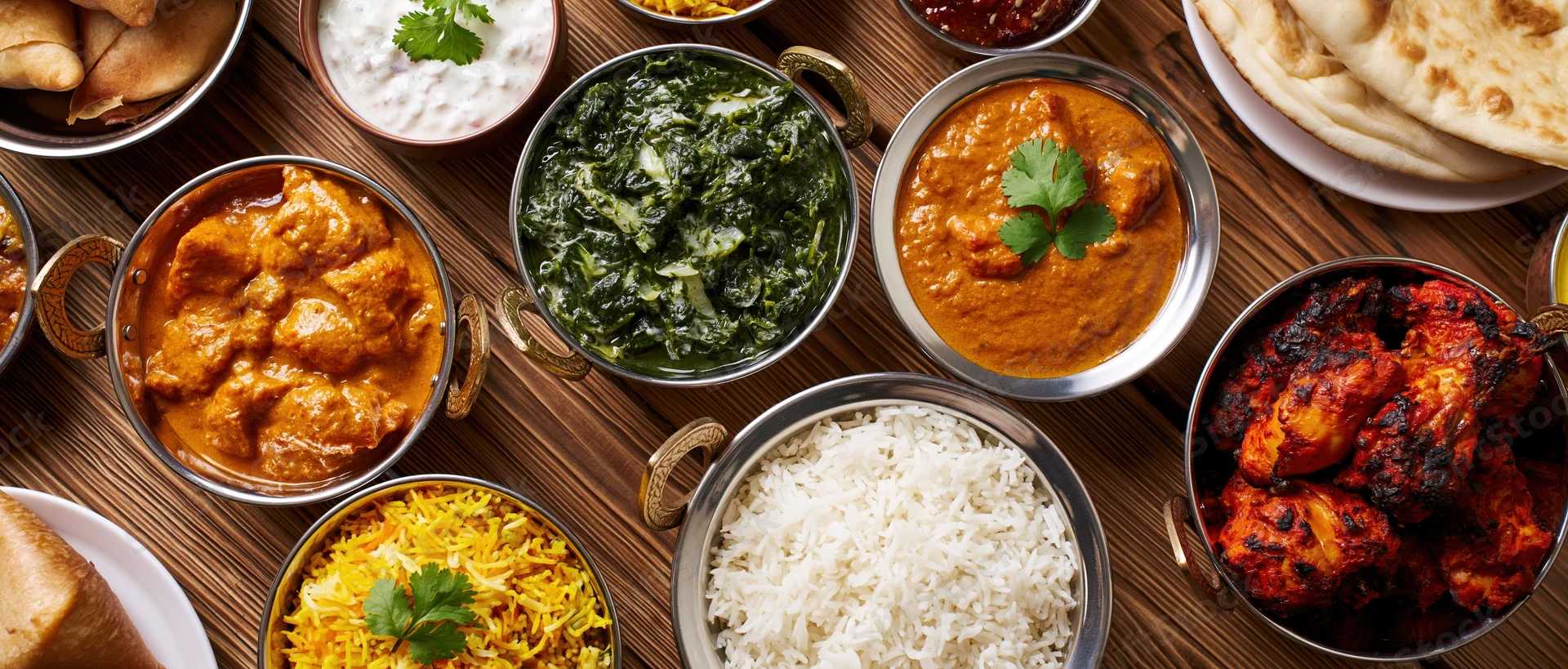 Overhead spread of indian food on wooden table with metal bowls and flatbreads