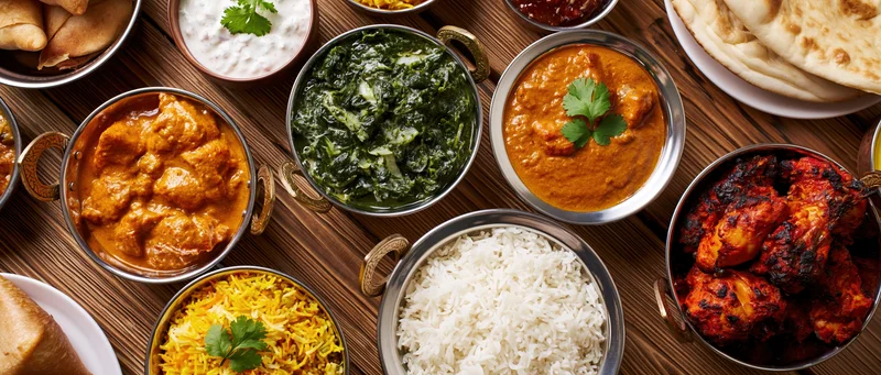 Overhead spread of indian food on wooden table with metal bowls and flatbreads