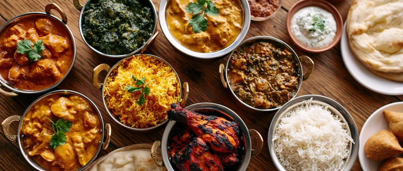 Overhead spread of indian dishes including curries, rice, naan and samosas on a wooden table