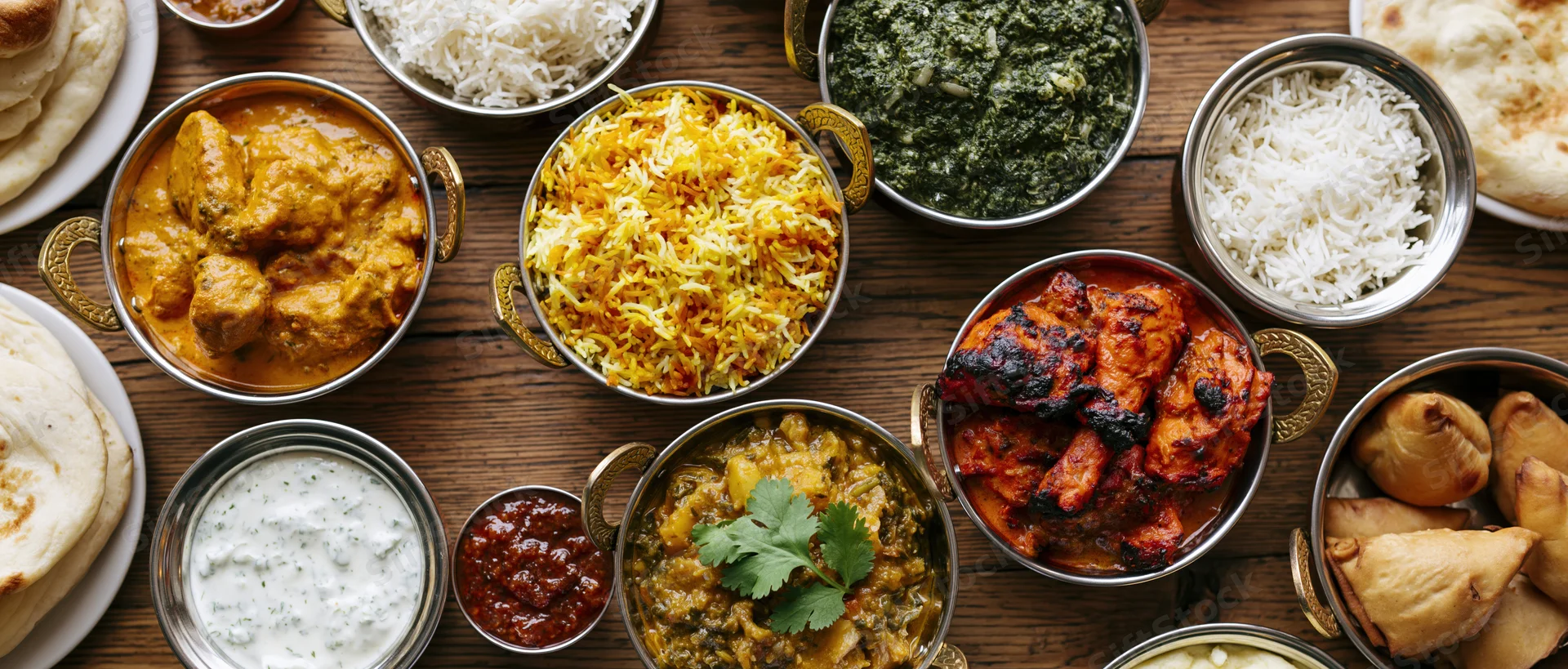 Overhead view of indian food spread on a worn wooden table with various curries, rice, naan, samosas, and chutneys arranged in metal bowls and plates