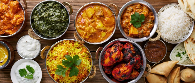 Overhead spread of indian food including curries, rice, naan, tandoori chicken, samosas, and raita on a wooden table