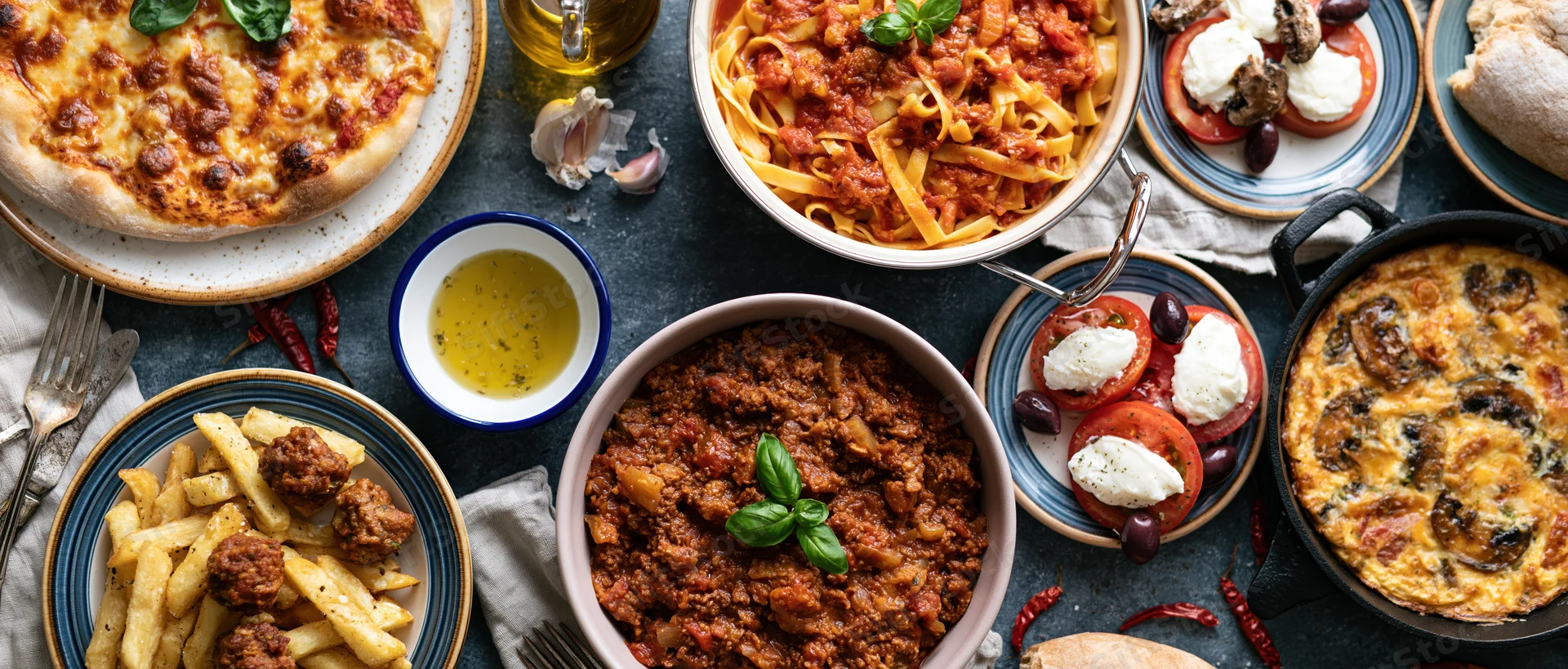 Overhead spread of italian dishes including pizza, pasta, caprese, and meatballs on a dark concrete table