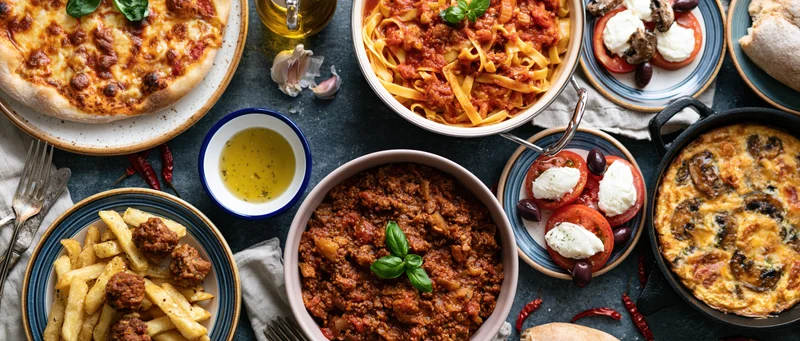 Overhead spread of italian dishes including pizza, pasta, caprese, and meatballs on a dark concrete table