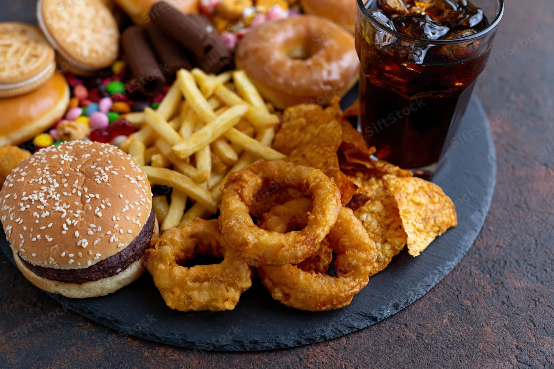 Overhead view of assorted junk food including burger, fries, onion rings, chips, soda, donuts, cookies and candy arranged on a dark slate board