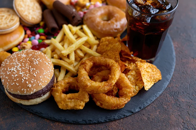 Overhead view of assorted junk food including burger, fries, onion rings, chips, soda, donuts, cookies and candy arranged on a dark slate board
