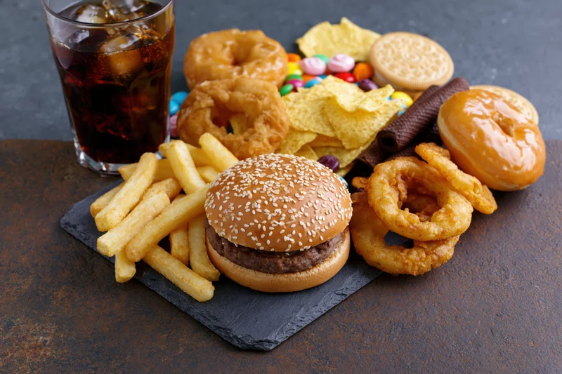 Overhead spread of junk food including burger, fries, soda, donuts, chips and candy arranged on dark slate board