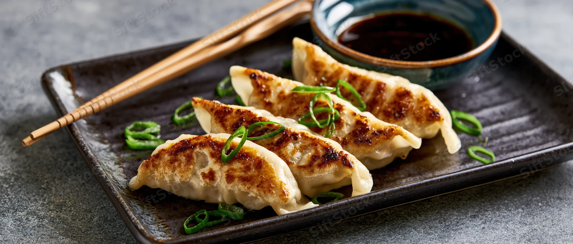 Pan-fried gyoza dumplings arranged in a row on a dark rectangular ceramic plate with chopsticks and soy sauce