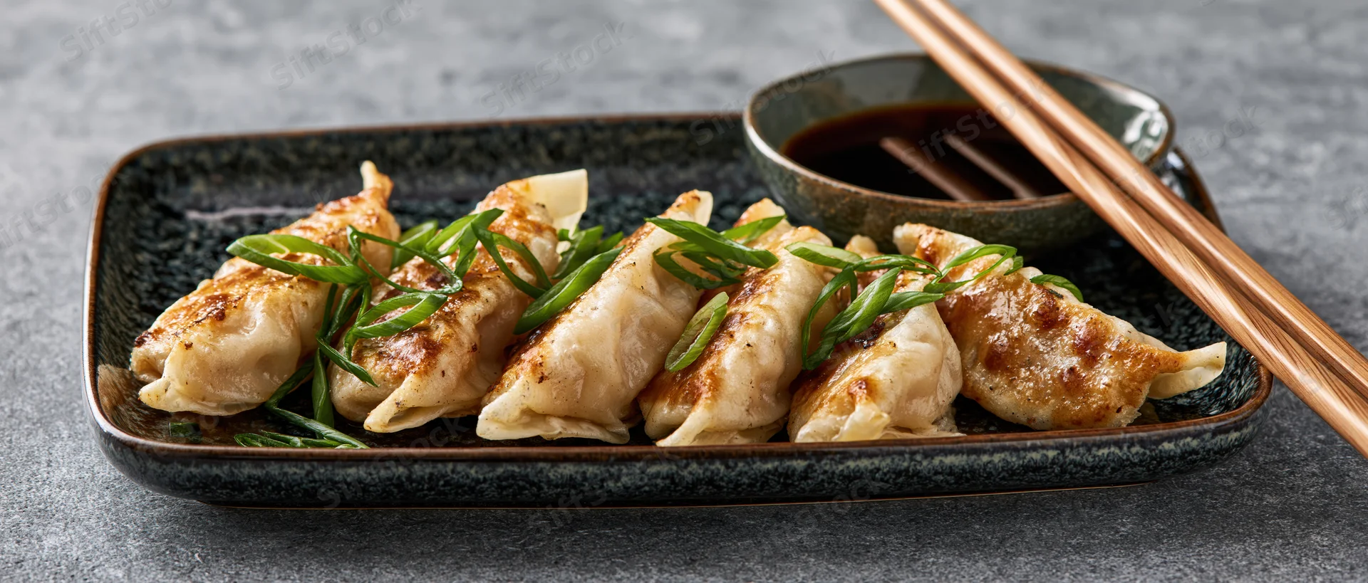 Pan-fried gyoza dumplings arranged in a row on a dark rectangular plate with soy sauce and chopsticks