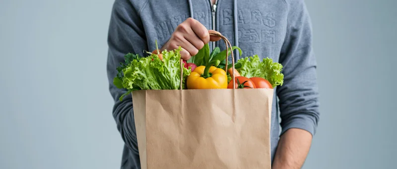 Man holding a brown paper bag filled with fresh vegetables against a plain background