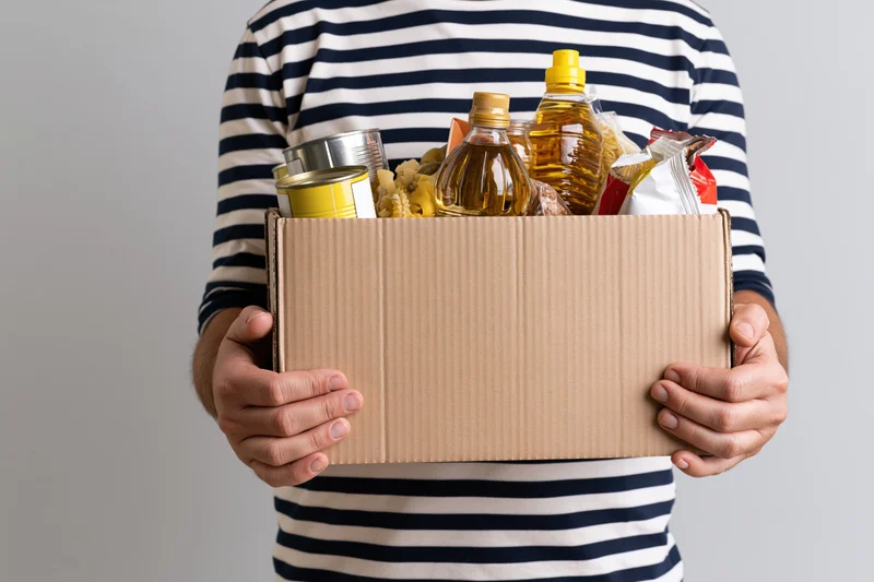 Man holding a cardboard box filled with canned goods and cooking oils against a gray background