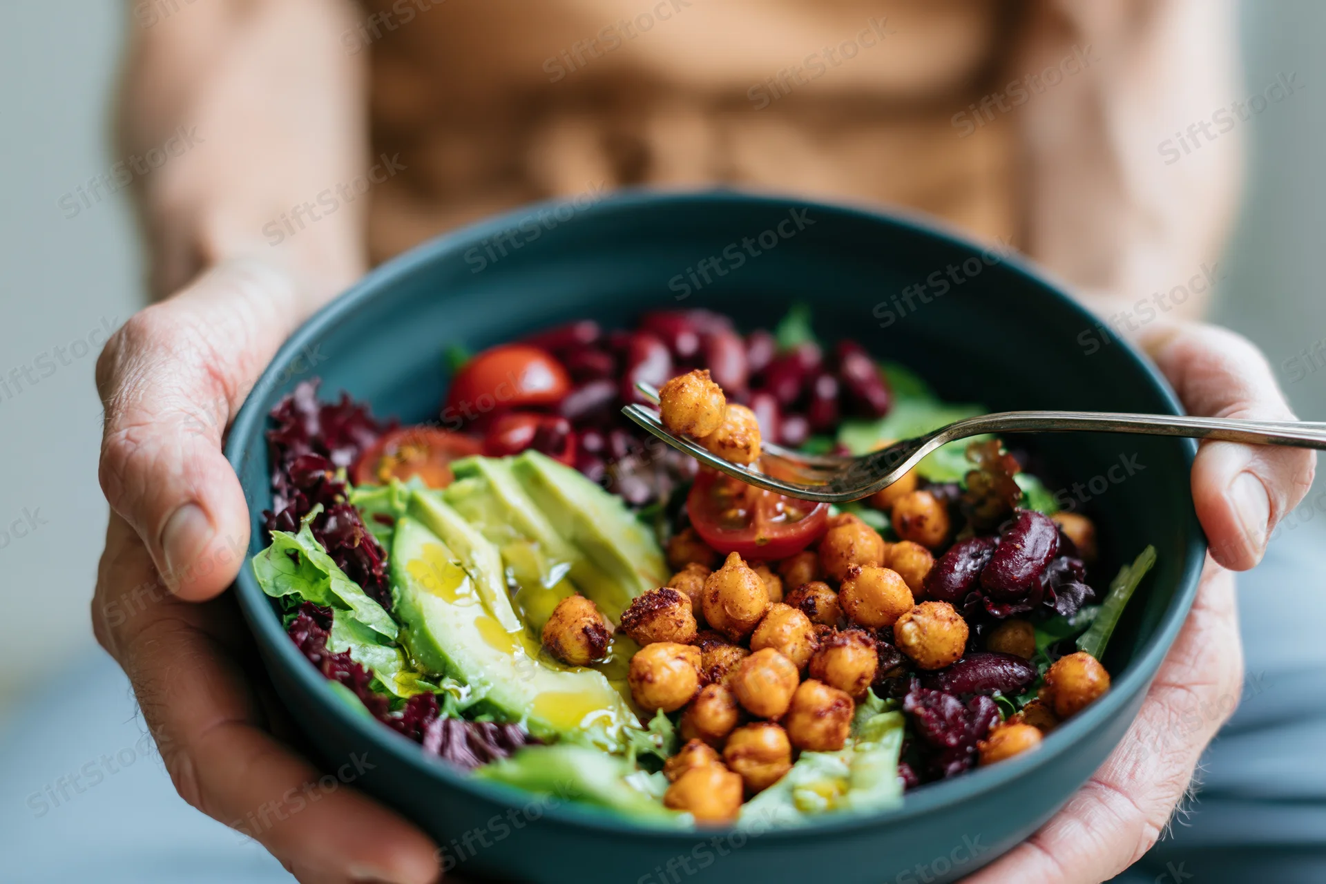 Hands holding a dark ceramic bowl filled with fresh salad of chickpeas, avocado, tomatoes, and lettuce